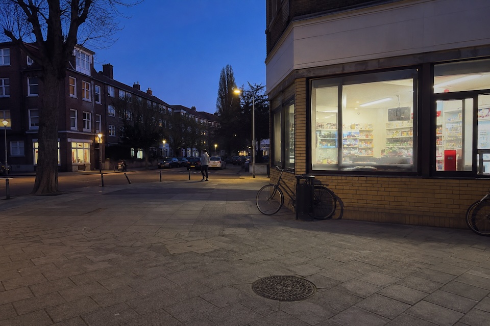 A lit corner shop, bicycles and a passerby at blue hour.