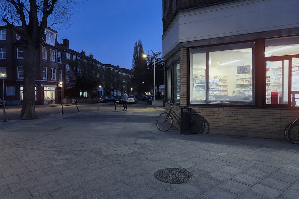 A lit corner shop, bicycles and a passerby at blue hour.