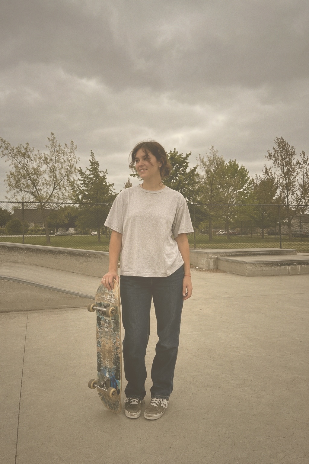A woman standing at a skatepark with a worn skateboard.